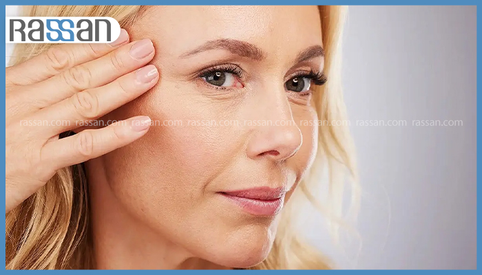 چطور روند پیری پوست صورت را کند کنیم؟ 5 زنی در حال بررسی خطوط ریز و نشانه های پیری روی پوست صورت و دور چشم خود با لمس دست